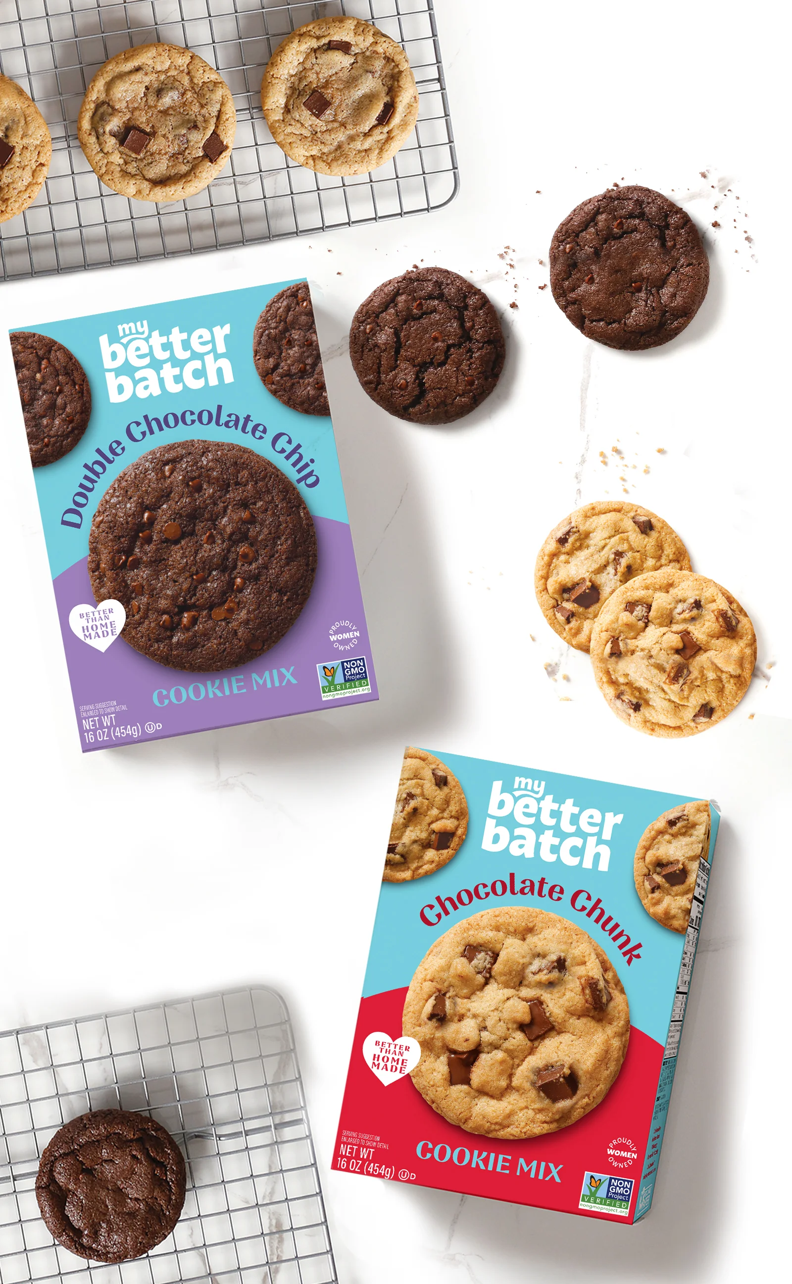 Two boxes of My Better Batch cookie mix on a white marble surface with cookies placed around them. The Double Chocolate Chip box is purple, and the Chocolate Chunk box is red. Both boxes display images of the cookies along with branding and certifications. Some cookies are cooling on a wire rack.